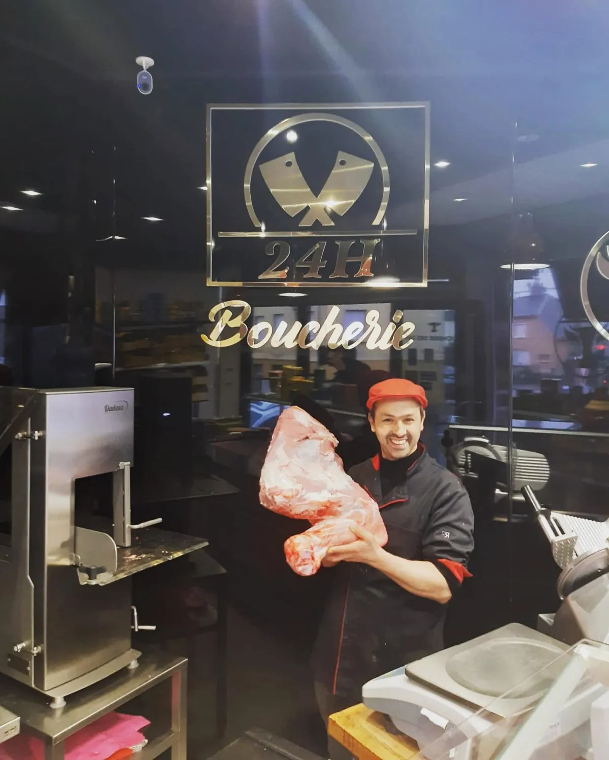 Le boucher derrière le comptoir, un gigot d'agneau dans les bras, sous l'enseigne dorée 24H Boucherie